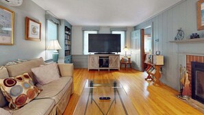 A living room with a sofa, television, bookshelf, and fireplace. - Cozy Cape style home near the beach and ferry with patio, W/D, & AC (Hyannis)