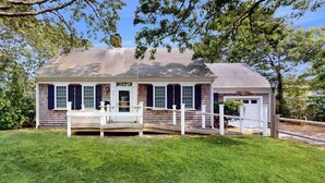 A single-story house with a porch, a small deck, and a well-maintained lawn. - Cozy Cape style home near the beach and ferry with patio, W/D, & AC (Hyannis)