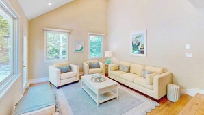 A spacious living room with two sofas, a coffee table, and a rug. There are windows with blinds, a lamp, and framed pictures on the walls. - Peaceful beach retreat: private yard, close to beaches, Chatham & rail trail (Harwich)