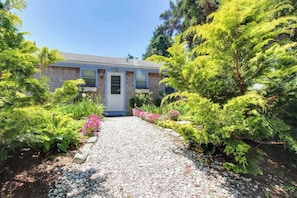 A house with a gravel driveway, surrounded by lush greenery and flowering plants. - Lakefront Cottage with a Deck, Free WiFi, Gas Grill - Close to Ferries & Beaches (Oak Bluffs)