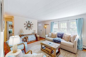 A living room with a sofa, coffee table, and a lamp. There is a window with curtains and a wall clock. - Conveniently-located home with fenced yard, firepit, beach parking pass (Hyannis)