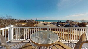 Een terras met een glazen tafel en stoelen, met uitzicht op een strand en een parkeerplaats met huizen op de achtergrond.