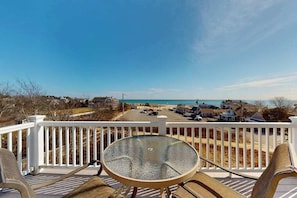 Eine Terrasse mit einem Glastisch und Stühlen, mit Blick auf einen Strand und einen Parkplatz mit Häusern im Hintergrund.