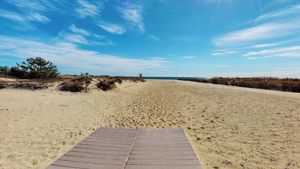 Een strand met een houten looppad naar het water, zandduinen en een verre redderstoren.
