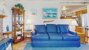 Ein Wohnzimmer mit einem blauen Sofa, einem Holzschrank mit Glastüren, einem Gemälde eines Leuchtturms und einem durch eine offene Tür einsehbaren Küchenbereich.