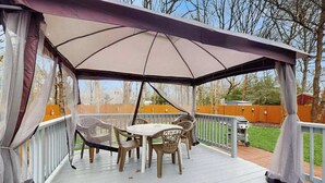 Eine überdachte Terrasse im Freien mit Tisch und Stühlen, einem Grill und Blick auf den Garten.
