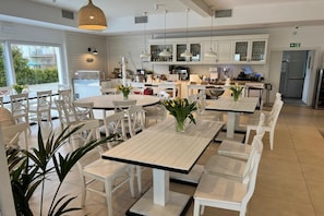 A modern dining area with white tables and chairs, a kitchen in the background, and a view of the outdoors. - Beautiful Apartment With a Terrace for 3 People (Krokowa)
