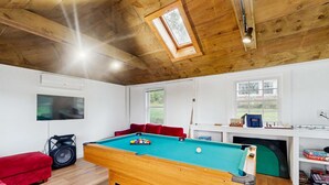 Een pooltafel in een ruimte met houten balkenplafond, een rode bank, een televisie en een raam.