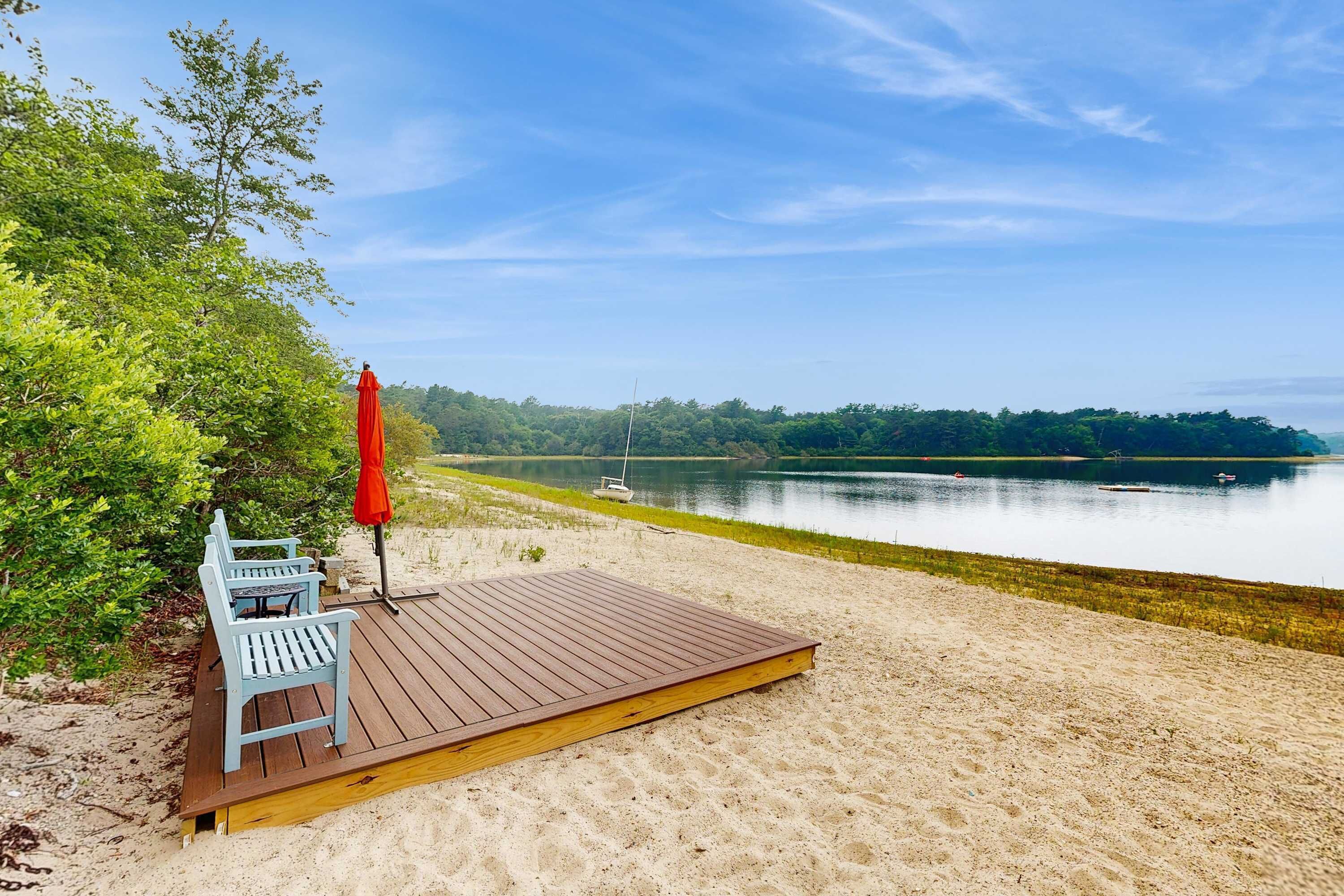 A wooden deck with a bench and a red umbrella, overlooking a calm lake with boats.