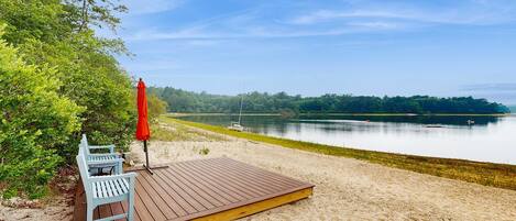 A wooden deck with a bench and a red umbrella, overlooking a calm lake with boats.
