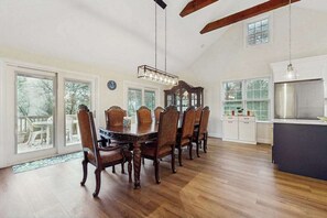 A spacious dining area with a large wooden table and chairs, a hanging light fixture, and a view through large windows. - Pond Treasure: Private beach, hot tub, sauna, play area & dog-friendly (Sandwich)