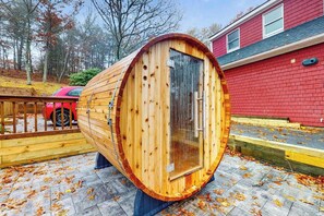 A wooden sauna with a glass door, set on a stone patio with fallen leaves. - Pond Treasure: Private beach, hot tub, sauna, play area & dog-friendly (Sandwich)