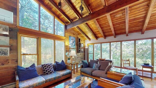 A log cabin interior with wooden beams, large windows, a sofa, and a coffee table. - Serene Home on 3 Acres w/ Wraparound Deck & Pool Table - Close to Beaches (Wellfleet)