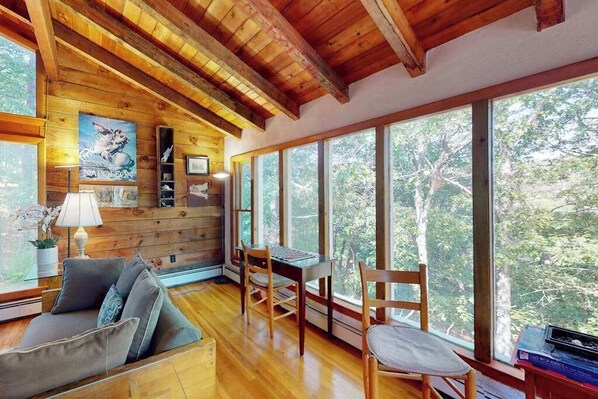 A wooden interior with a sofa, chairs, a table, and large windows offering a forest view. - Serene Home on 3 Acres w/ Wraparound Deck & Pool Table - Close to Beaches (Wellfleet)