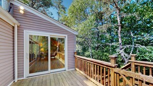 A wooden deck with a glass door leading to a covered outdoor area, surrounded by trees. - Serene Home on 3 Acres w/ Wraparound Deck & Pool Table - Close to Beaches (Wellfleet)