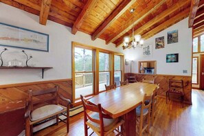 A wooden dining area with a large table and chairs, wooden beams on the ceiling, and large windows. - Serene Home on 3 Acres w/ Wraparound Deck & Pool Table - Close to Beaches (Wellfleet)