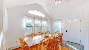 Ein Esszimmer mit Holztisch und -stühlen, großen Fenstern mit Vorhängen, einem Deckenventilator und einer weißen Tür.