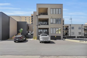 Ein modernes Gebäude mit Parkplatz, zwei abgestellten Fahrzeugen und einer Treppe zum Eingang.