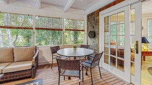 A covered patio with a round table and chairs, a sofa, and a view through the windows. - Beautiful Estate with Large Lawn Made for Socializing and Kids' Playroom (Vineyard Haven)