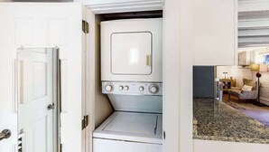 A compact laundry room with a washer and dryer, a door leading to a living area with a sofa and lamp. - Modern & bright condo with patio, W/D, and central location near town & beach (Provincetown)
