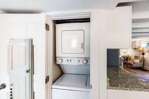 A compact laundry room with a washer and dryer, a door leading to a living area with a sofa and lamp. - Modern & bright condo with patio, W/D, and central location near town & beach (Provincetown)