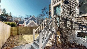 A backyard with a wooden fence, a concrete staircase, and a house with a gabled roof. - Modern & bright condo with patio, W/D, and central location near town & beach (Provincetown)