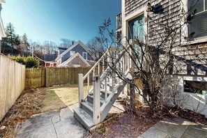 A backyard with a wooden fence, a concrete staircase, and a house with a gabled roof. - Modern & bright condo with patio, W/D, and central location near town & beach (Provincetown)