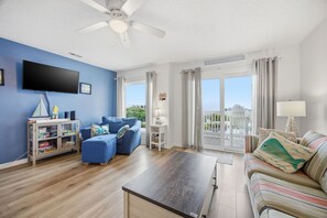 Un salón con televisor de pantalla plana, sofá azul, sofá a rayas, mesa de centro de madera y ventilador de techo.