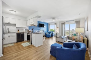 A modern kitchen with white cabinets, a central island, and a dining area with a blue sofa and a small table. - Short Way to the Beach - Free $100 Grocery Gift Card (Emerald Isle)