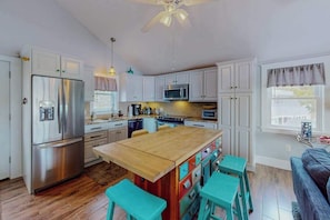 Una cocina con gabinetes blancos, electrodomésticos de acero inoxidable, una isla de madera con taburetes y un ventilador de techo.