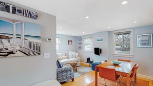 Uma sala de estar com vista para o oceano, uma área de jantar com mesa e cadeiras de madeira, um sofá branco e uma poltrona estampada.