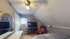 Un dormitorio con cama, televisor, ventilador de techo, silla y cómoda.