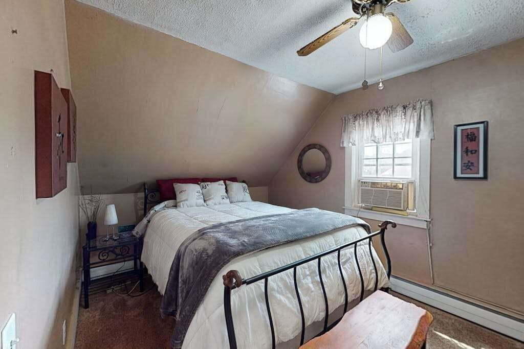 A bedroom with a bed, a bedside table, a lamp, a ceiling fan, a window with curtains, and a wall-mounted picture frame.