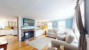 Ein Wohnzimmer mit Kamin, einer Couch, einem Fernseher und einer Küchenzeile mit Schränken und einer Arbeitsplatte.