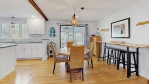 Une cuisine moderne avec des meubles blancs, une table à manger avec des chaises, un luminaire suspendu et une fenêtre avec des rideaux.