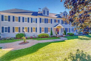 A large, two-story house with a symmetrical facade, a central entrance with a covered porch, and a well-maintained lawn. - Sprawling dog-friendly lake estate with pool, patio, fire pit, games, & views (Barnstable)