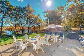 An outdoor patio with Adirondack chairs, a round table, and a grill area. - Sprawling dog-friendly lake estate with pool, patio, fire pit, games, & views (Barnstable)