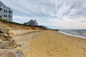 A beach with a house on a rocky cliff, a wooden walkway, and the ocean. - Dreamy ocean-view perch with furnished balcony & beach access - dog-friendly (Dennis Port)