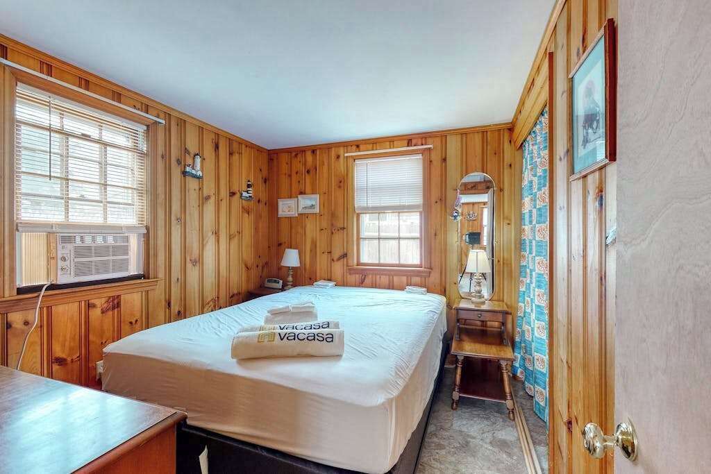 Habitación con paneles de madera, una cama, mesita de noche, lámpara y una ventana con persianas.