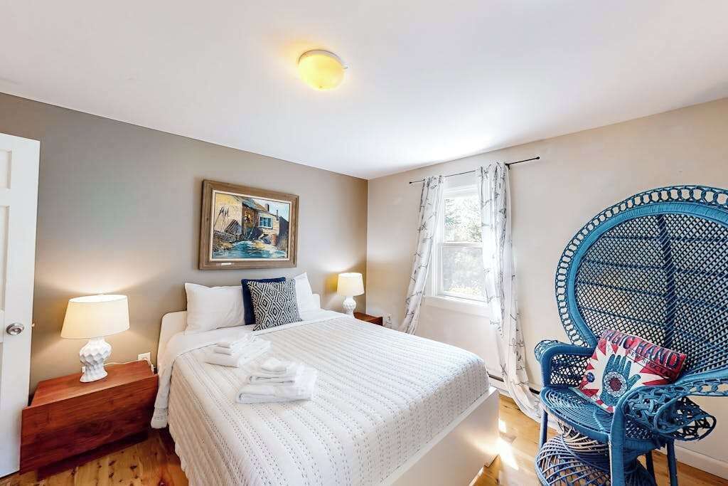 A bedroom with a bed, two bedside lamps, a framed picture, a window with curtains, and a blue chair with a magazine.