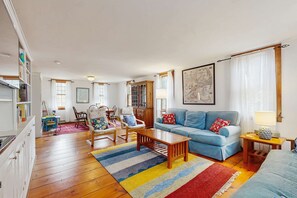 Un salon avec un canapé bleu, une table basse en bois et un tapis à rayures. On y trouve également un coin repas avec des chaises et une table, ainsi qu’une cuisine équipée de placards et d’un réfrigérateur.