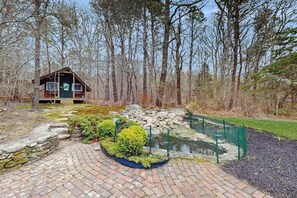 Une cabane avec un sentier de pierres menant à un étang, le tout entouré d’arbres.