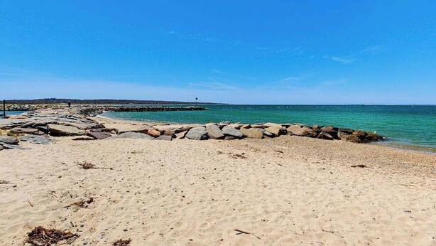 Ein Strand mit einem steinernen Wellenbrecher, blauem Himmel und ruhiger See.