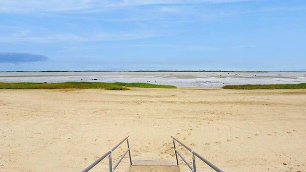 Uma praia com uma escadaria que leva a uma área arenosa, céu claro e mar calmo.