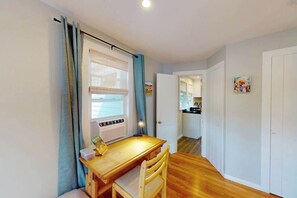 A room with a wooden desk, a chair, a window with curtains, a door, and a hallway leading to a kitchen. - Cozy home close to the beach with patio, Washer/Dryer, & Partial AC (Barnstable)