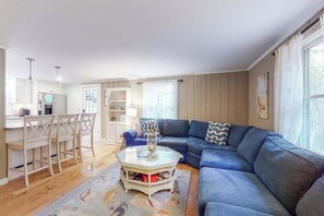 Ein Wohnzimmer mit blauem Sofa, einem Couchtisch und einem Essbereich mit Stühlen, sowie einer Küche im Hintergrund.