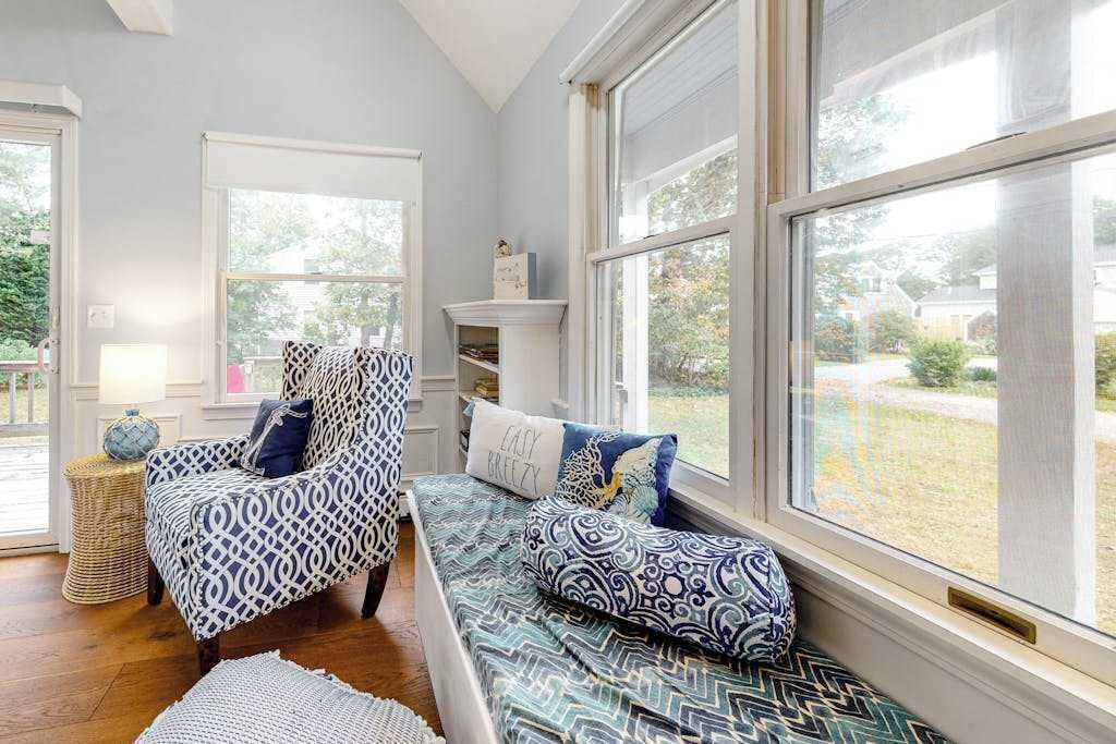 A room with a large window, a patterned chair, a bench with cushions, and a small table with a lamp.