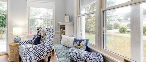 A room with a large window, a patterned chair, a bench with cushions, and a small table with a lamp.