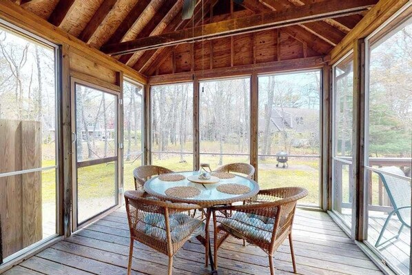 A covered outdoor area with a round table and wicker chairs, large windows, and a wooden ceiling. - Peaceful cottage with deck, screened porch, grill, & firepit - walk to beach (Eastham)