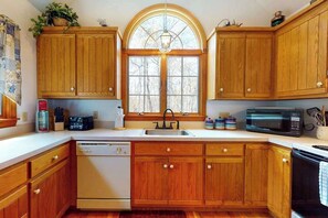 A kitchen with wooden cabinets, a dishwasher, a microwave, and a window with a view of trees. - Peaceful cottage with deck, screened porch, grill, & firepit - walk to beach (Eastham)